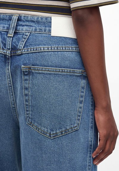 Personne portant un jean bleu en denim et une chemise à rayures, vue de la taille vers le bas, avec une poche arrière visible et la main sur le côté.