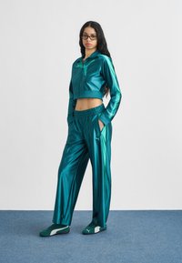 Γυναίκα με μακριά σκουρόχρωμα μαλλιά, που φορά γυαλιά και γυαλιστερή φόρμα Puma σε τιρκουάζ χρώμα, στέκεται σε μπλε χαλί μπροστά σε λευκό φόντο.