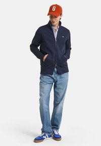 Giacca zip-up blu navy con colletto alto, due tasche laterali, indossata sopra una camicia a scacchi. Abbinata a jeans azzurri e sneakers blu.