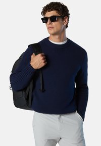 Pull en maille bleu marine avec des manches et un ourlet côtelés, porté par un mannequin tenant un sac à dos noir. Le pull a un col rond.