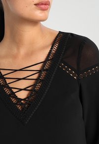 Zwarte blouse met een V-hals, voorzien van kantdetails en kruislingse bandjes. Doorzichtige stof op de schouders voegt textuur en contrast toe.