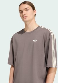 Sivá tričko s krátkym rukávom a voľným strihom, s bielymi tromi pruhmi na ramenách a malým bielym logom na hrudi.