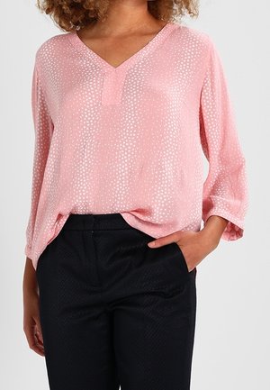 Femme portant un chemisier rose clair à manches longues avec des pois blancs et un pantalon noir texturé, une main dans la poche.