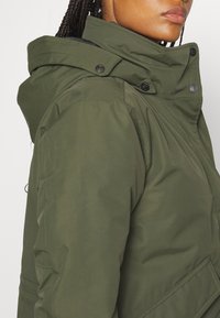 Olivovo zelená bomber bunda s vysokým golierom, so zapínaním na cvoky a štruktúrovaným povrchom. S kapucňou a bočnými vreckami.
