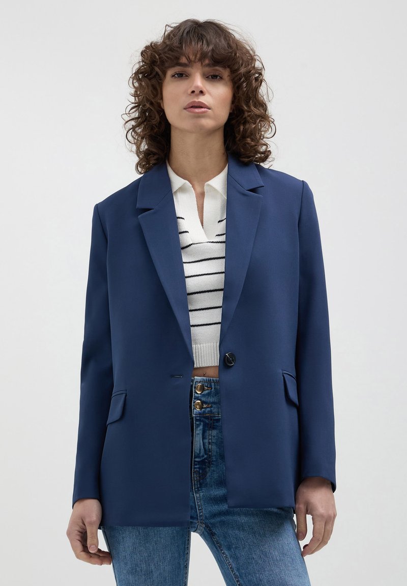 Femme aux cheveux bouclés portant un blazer bleu marine sur un top en maille blanche rayé et un jean bleu taille haute avec des détails de boutons.