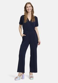 Marineblauwe jumpsuit met korte mouwen, wikkeldesign, strik taille en zakken. Gemaakt met metallic platform sandalen. Zachte stof textuur.