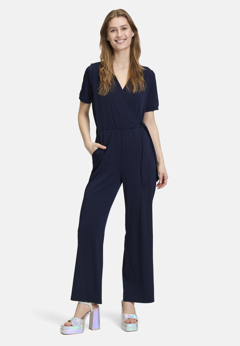 Marineblauwe jumpsuit met korte mouwen, wikkeldesign, strik taille en zakken. Gemaakt met metallic platform sandalen. Zachte stof textuur.