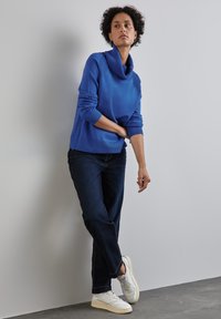 Blauer, grob gestrickter Pullover mit einem Schalkragen, kombiniert mit dunkelblauen Jeans und weißen Sneakern, die eine lässige Passform und strukturierte Details hervorheben.