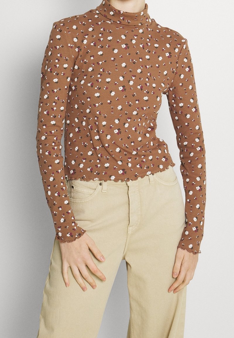 Braunes, floral gemustertes Langarmoberteil mit Stehkragen, das kleine weiße und rote Blumen sowie eine gerippte Textur und einen volantsaum aufweist. Kombiniert mit beigen Hosen.