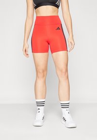 Punased sportlikud lühikesed püksid kõrge vöökohaga, mustade külgtriipudega ja Adidas logo. Kombineeritud valgete tossude ja triibuliste sokkidega.