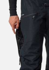 Pantalons de bavoir noirs imperméables avec poche zippée sur le côté, coutures renforcées et bretelles ajustables. Le matériau semble lisse avec un léger éclat.