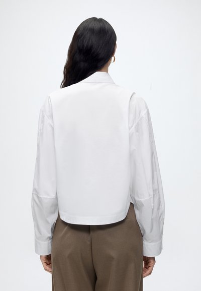 Femme aux longs cheveux foncés portant une chemise blanche à manches longues et un pantalon marron, debout de dos devant un fond clair uni.