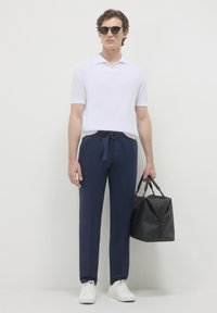 Weißes Polo-Shirt mit kurzen Ärmeln, marineblaue Hose mit Gürtel, schwarze Tasche und weiße Sneaker. Modell trägt schwarze Sonnenbrille.