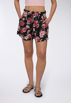 Pantaloni corti neri a fiori con rose rosa e bianche, vita elastica con cordino, tasche laterali, tessuto leggero, abbinati a sandali con le stringhe.