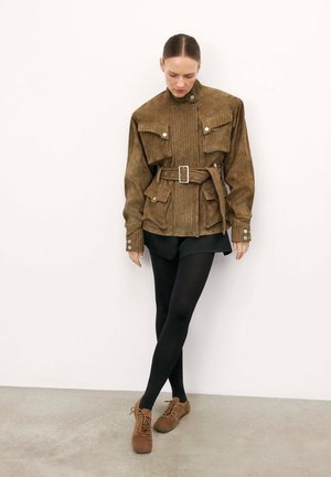 Bruine suède oversized jasje met een hoge kraag, borstzakken, een ceintuur in de taille en zilverkleurige accenten, gecombineerd met zwarte shorts en maillot.