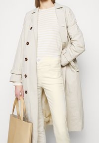 Licht beige trenchcoat met grote knopen, gelaagd over een crèmekleurig en lichtgeel gestreept shirt, gecombineerd met crèmekleurige broek en een tan schoudertas.
