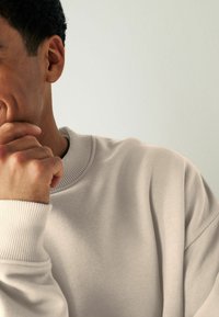 Sweatshirt beige clair avec col et poignets côtelés, au toucher doux. Vue de côté montrant le bras et la main près du menton, sur un fond doux.