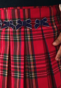 Röd tartankjol med marinblå sammetssnören på midjebandet. Tyget har veck och en blandning av gröna, gula och vita ränder.