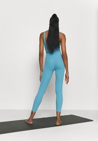 Blaues sportliches Crop-Top und Leggings, figurbetontes Design, glatte Textur. Stehend auf einer dunklen Yogamatte, von hinten gesehen, mit langen schwarzen Haaren.