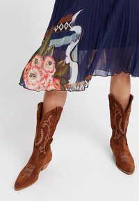 Stivali da cowboy in suede marrone con punte rialzate e ricami, abbinati a una gonna plissettata blu con vivaci disegni floreali.