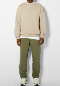 Beige sweater met ronde hals en "PARIS" geborduurd op de borst, gecombineerd met groene cargobroek en crèmekleurige sneakers voor een ontspannen look.