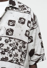 Gros plan d'une chemise à manches courtes avec des motifs floraux et de feuilles noirs sur un fond gris clair.