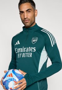 Grøn langærmet atletisk top med høj krave, hvide detaljer og et Arsenal-logo. Holder en blå og hvid mønstret fodbold.