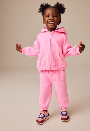 Lächendes Kleinkind mit lockigem Haar in rosa Kapuzen-Trainingsanzug und bunten Turnschuhen steht mit leicht erhobenen Armen vor einfachem Hintergrund.