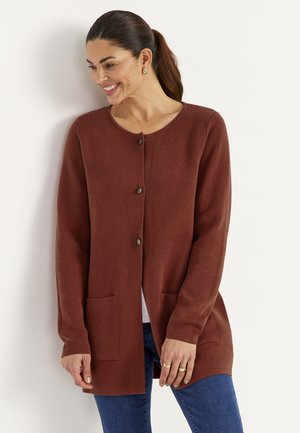 Frau lächelt, trägt eine braune, geknöpfte Strickjacke mit Fronttaschen, blaue Jeans und goldene Creolen vor weißem Hintergrund.