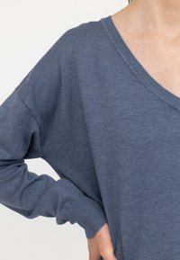 Maglione blu lavorato a maglia con scollatura a V ampia, spalle drop e polsini a coste. La texture del tessuto appare morbida e leggera.