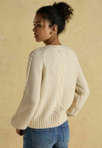 Svetre v krémovej farbe s pleteným vzorom, rebrovaným lemom a zložitým diamantovým dizajnom na chrbte. Rukávy sú vo voľnom strihu.