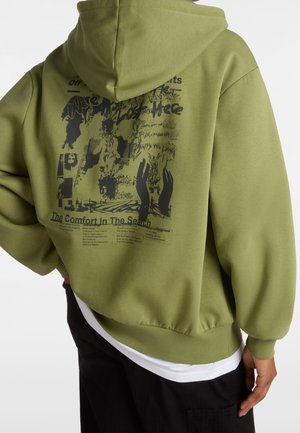 Personne portant un sweat à capuche vert olive avec un graphisme abstrait noir et du texte dans le dos, associé à un pantalon noir et une chemise blanche en dessous.