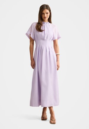 Kvinde står i en gulvlang lavendelfarvet kjole med plisseringer og flagrende ærmer, iført åbne hæle og et armbånd på højre håndled.