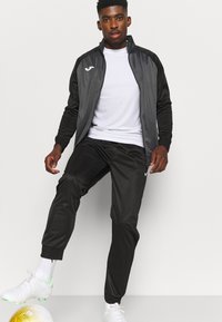 Zwarte sportjack met grijze accenten, wit logo op de borst, gecombineerd met zwarte broek. Model draagt een wit shirt en stijlvolle witte sportschoenen.