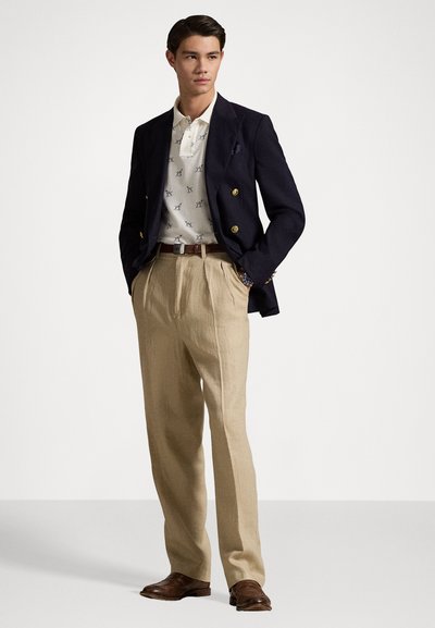 Marineblazer met gouden knopen, crèmekleurige polo met sterrenpatroon, beige geplooide broek, bruin lederen schoenen en een brown riem.
