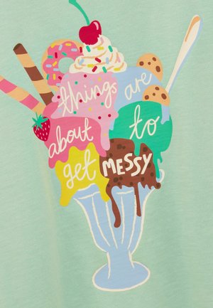 Eisbecher mit fünf Kugeln, Schlagsahne, Kirsche, Keksen, Keksstäbchen und dem Text „Jetzt wird es chaotisch“ vor hellgrünem Hintergrund.