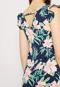 Květinové šaty s námořnickým pozadím, zdobené růžovými hibisky a zelenými listy, mají krátké volánkové rukávy a V-otvor na zádech.