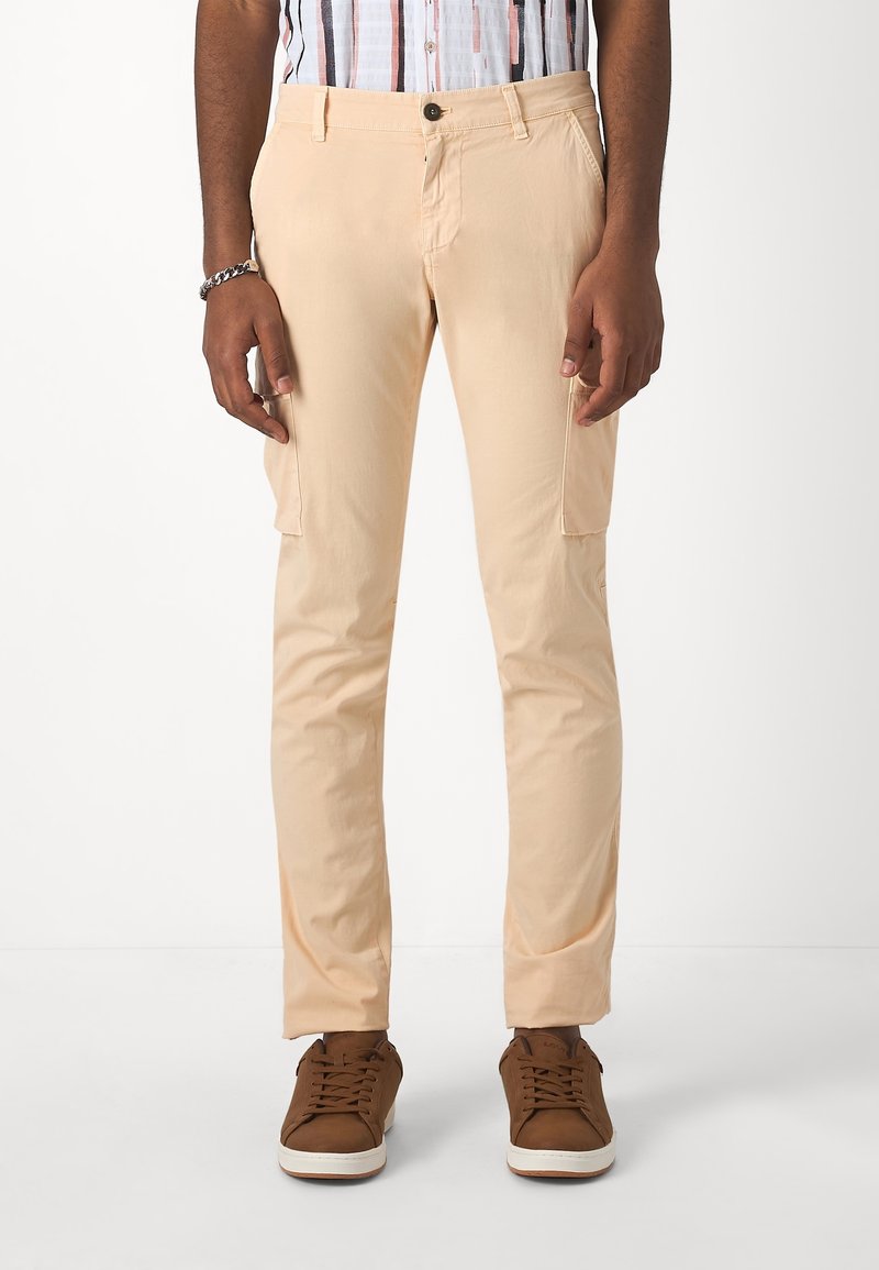Lichtbeige cargobroek met twee zijzakken, knoopsluiting aan de voorkant en een taps toelopende pasvorm, gecombineerd met bruine sneakers.