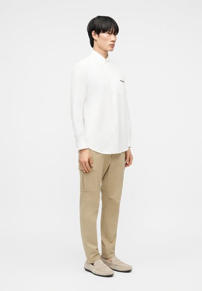 Άνδρας όρθιος, φοράει λευκό πουκάμισο με κουμπιά, μπεζ παντελόνι cargo και μπεζ παπούτσια slip-on, πάνω σε απλό λευκό φόντο.