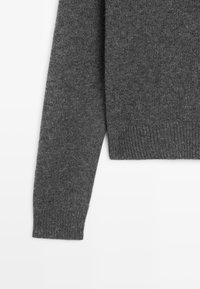 Un maglione grigio a maglia con scollo rotondo e orlo a coste. Il tessuto ha una consistenza morbida e una vestibilità leggermente ampia.