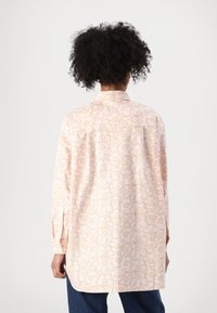 Camisa de manga larga en tejido rosa claro con un estampado floral en dorado. Presenta un cuello y un dobladillo redondeado, con un corte relajado.