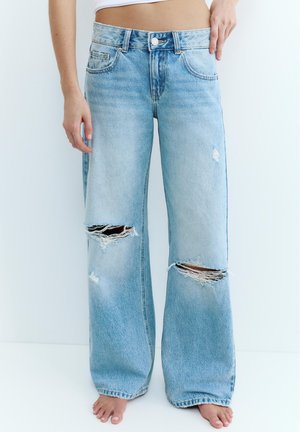 Hellblaue Jeans mit weitem Bein und aufgerissenen Stellen an beiden Knien, getragen von einer barfüßigen Person vor weißem Hintergrund.