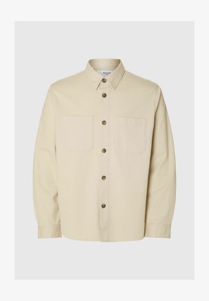 Beige košile s dlouhým rukávem na knoflíky, se dvěma kapsami na prsou a hnědými knoflíky, vystavená na jednolitém pozadí.