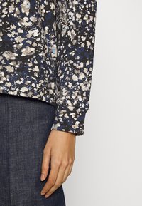 Käsi puhkab külje peal, kandes tumedat sinist täpilist pikka varrukat ja tumedaid teksaseid valge tausta taustal.