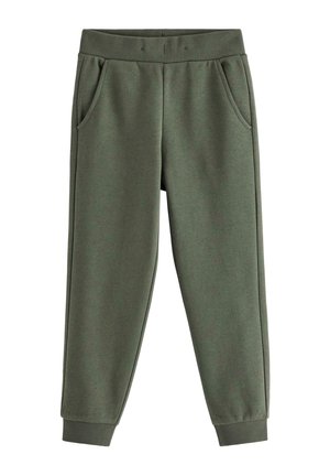 Ung dreng står og smiler, iført en beige stribet langærmet skjorte, mørkegrønne bukser og lyse sneakers med blå stropper.
