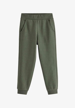 Ung dreng står og smiler, iført en beige stribet langærmet skjorte, mørkegrønne bukser og lyse sneakers med blå stropper.