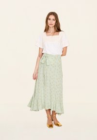 Weißes Oberteil mit bestickten Details, kombiniert mit einem mintgrünen, floral gemusterten Wickelrock. Das Outfit hat eine fließende Silhouette und goldene Schuhe.