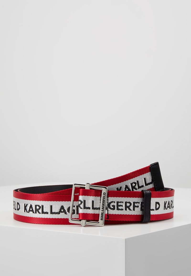 Ceinture en tissu rouge et blanc avec texte noir "KARL LAGERFELD" et boucle argentée exposée sur une surface blanche.