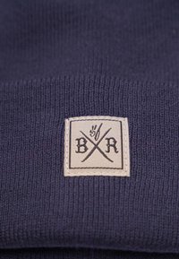 Laine en tricot bleu marine avec une étiquette en tissu portant "BXR" et des axes croisés dans un carré. Texture douce avec un design en côtes simple.