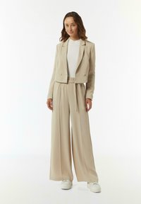 Beige op maat gemaakte blazer met dubbele knopen, gecombineerd met beige wijde broeken met een elastische tailleband. Witte sneakers maken de outfit compleet.